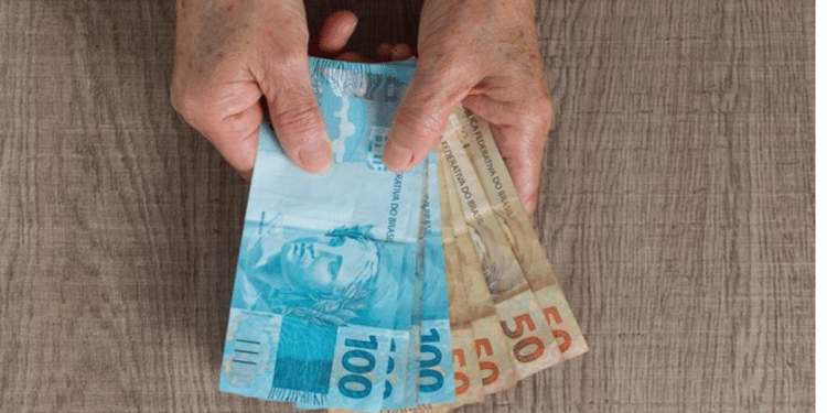 Mãos de pessoa idosa segurando notas de 100 e 50 reais sobre mesa de madeira.