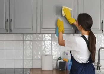 Mulher usando luvas amarelas limpando armário de cozinha cinza com pano branco