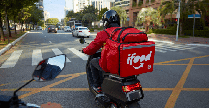 Motociclista entregador do iFood com mochila térmica vermelha trafegando em avenida movimentada