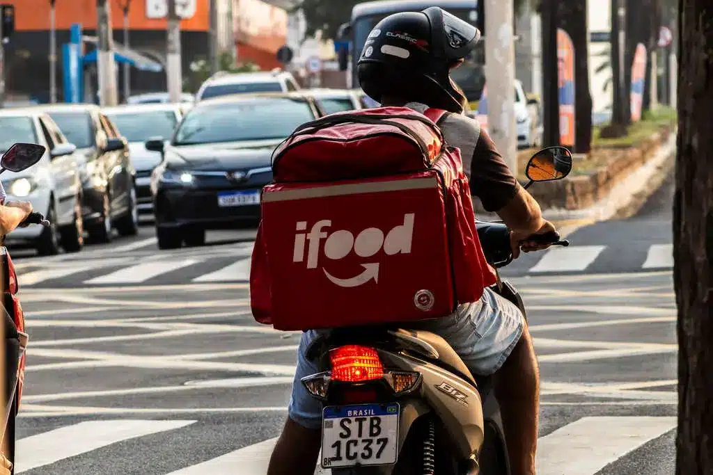 Entregadores iFood Entregador de aplicativo circulando de moto em área urbana, representando critérios de elegibilidade para bônus por cidade.