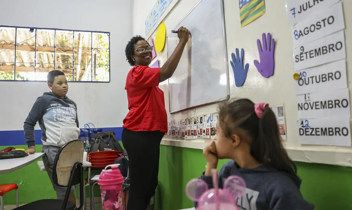 Professora ensinando alunos em sala de aula colorida Professora sorridente escrevendo no quadro branco com dois alunos observando atentos em sala de aula decorada