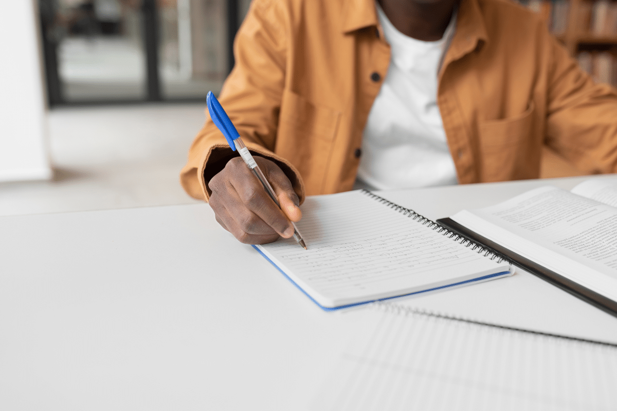 Pessoa negra com camisa marrom escreve em caderno espiral aberto na mesa branca com livro ao lado