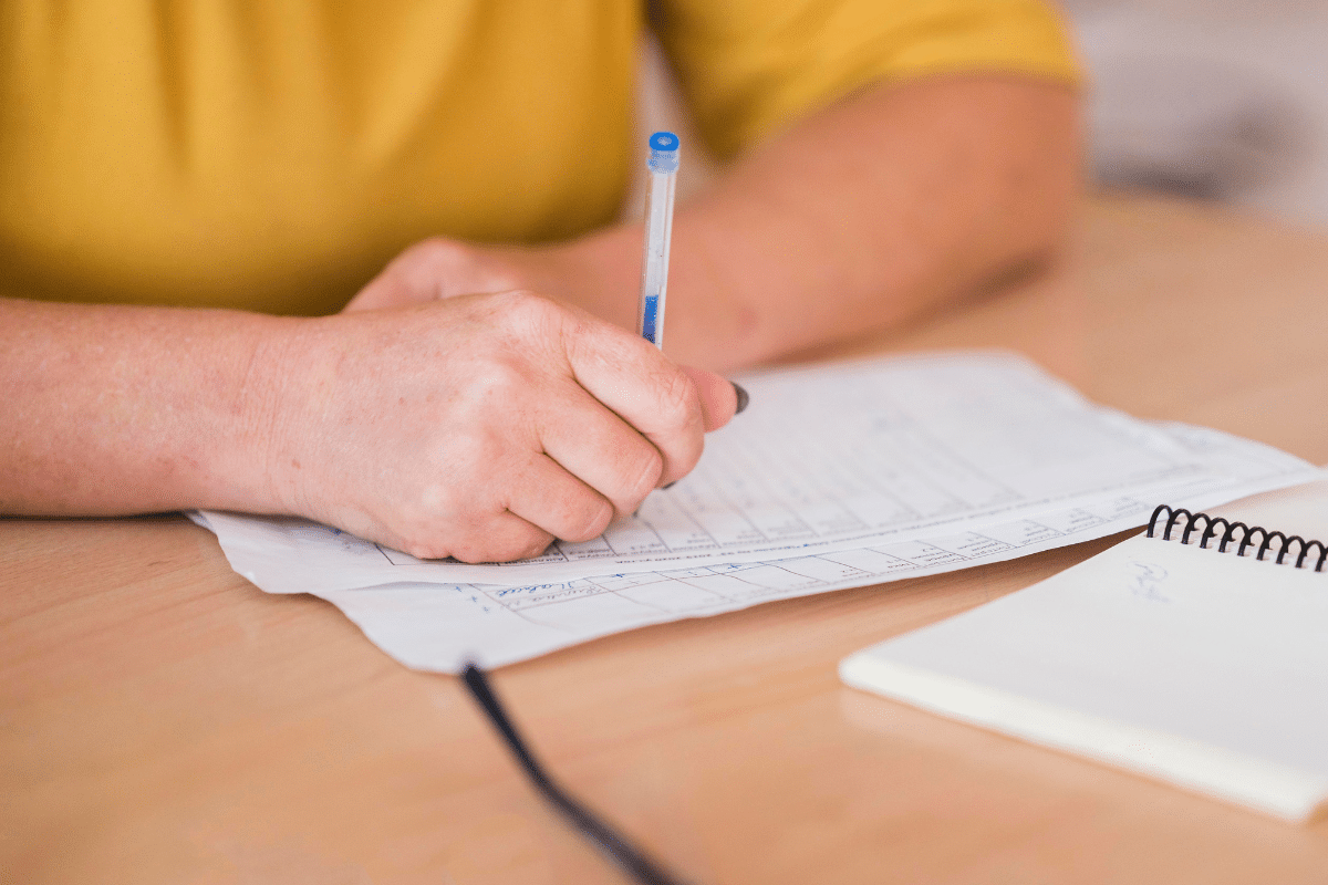 Mão escrevendo em formulário com caneta azul sobre mesa de madeira clara, próximo a caderno espiral