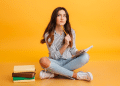 Mulher jovem sentada no chão com caderno e lápis, pensando, com pilha de livros ao lado e fundo amarelo