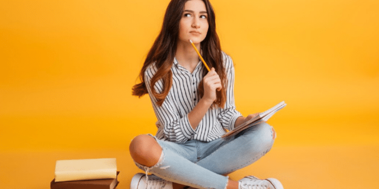 Mulher jovem sentada no chão com caderno e lápis, pensando, com pilha de livros ao lado e fundo amarelo