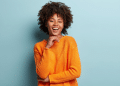 Mulher negra jovem com cabelo afro usando suéter laranja sorrindo com fundo azul claro