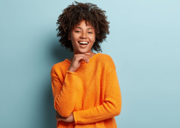Mulher negra jovem com cabelo afro usando suéter laranja sorrindo com fundo azul claro