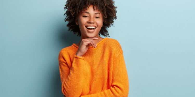 Mulher negra jovem com cabelo afro usando suéter laranja sorrindo com fundo azul claro