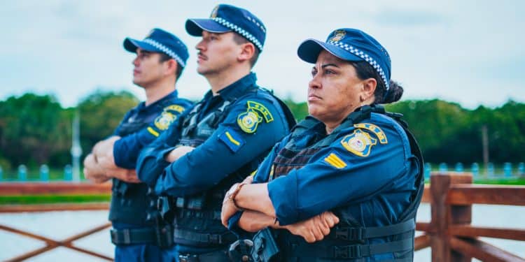 Três guardas municipais com coletes e uniformes azul-marinho olhando à frente com braços cruzados em área externa