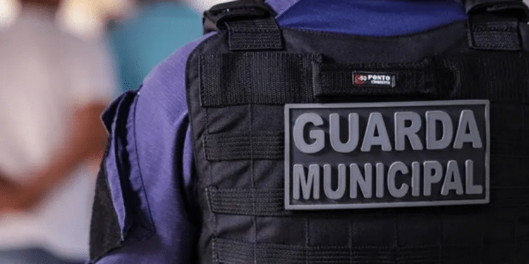 Guarda Municipal de costas usando colete tático preto e uniforme azul em ambiente desfocado.