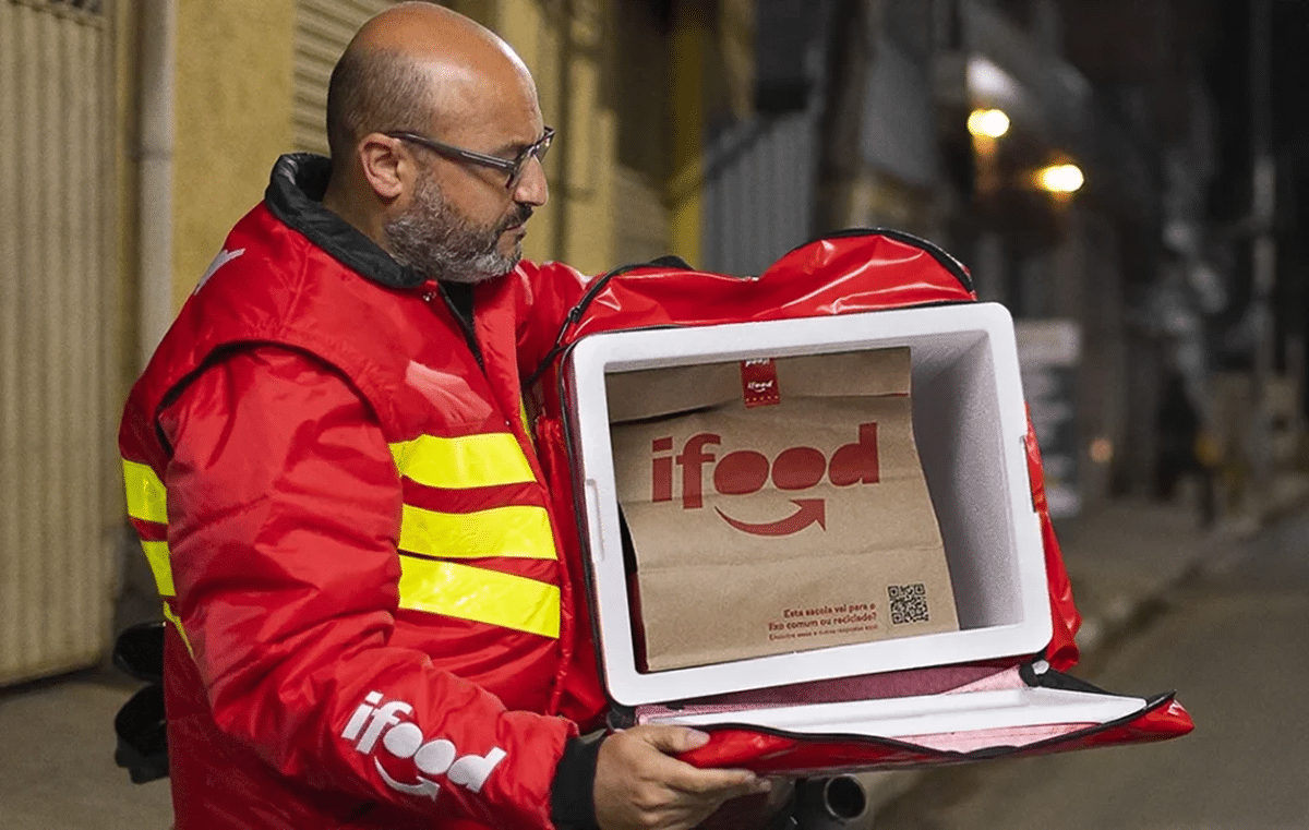 Entregador do iFood vestindo jaqueta vermelha com faixas refletivas retirando pedido da bag térmica durante entrega noturna.