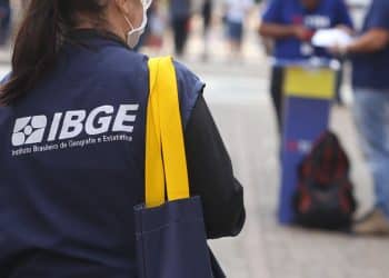 Mulher com colete do IBGE e máscara, carregando bolsa azul com alça amarela em área urbana