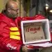 Homem usando jaqueta vermelha de entregador iFood segurando mochila térmica aberta com embalagem de papel iFood