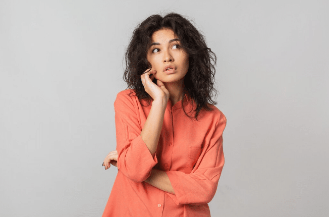 Mulher jovem pensativa vestindo camisa laranja olhando para cima com fundo cinza