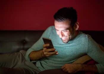 Homem sorrindo e relaxado, usando o smartphone no sofá durante a noite, representando o consumo viciante de conteúdo, como os Reels do Instagram, na madrugada.