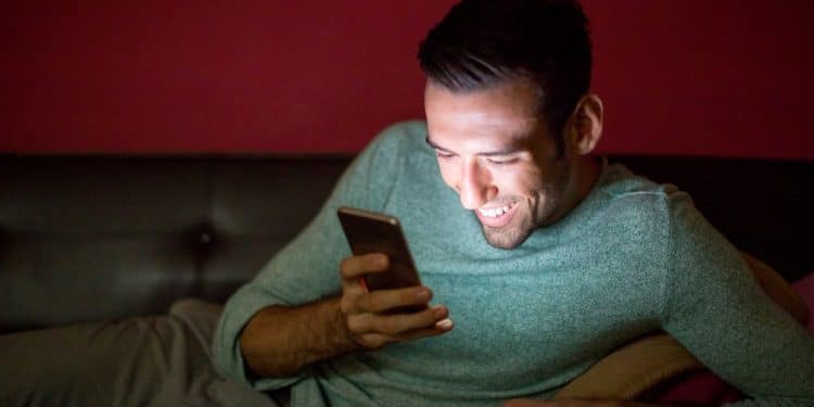 Homem sorrindo e relaxado, usando o smartphone no sofá durante a noite, representando o consumo viciante de conteúdo, como os Reels do Instagram, na madrugada.