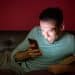 Homem sorrindo e relaxado, usando o smartphone no sofá durante a noite, representando o consumo viciante de conteúdo, como os Reels do Instagram, na madrugada.