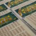 Close de vários bilhetes oficiais da Mega da Virada espalhados sobre uma mesa, exibindo o logotipo dourado do sorteio e as tabelas de números para marcação.