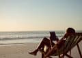 Vista lateral de uma pessoa sentada em uma cadeira de praia listrada na areia, usando um tablet enquanto observa o mar sob a luz suave do entardecer.