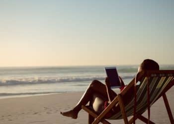 Vista lateral de uma pessoa sentada em uma cadeira de praia listrada na areia, usando um tablet enquanto observa o mar sob a luz suave do entardecer.