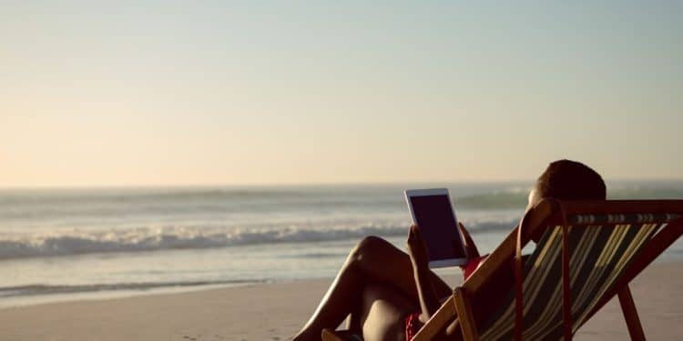 Vista lateral de uma pessoa sentada em uma cadeira de praia listrada na areia, usando um tablet enquanto observa o mar sob a luz suave do entardecer.