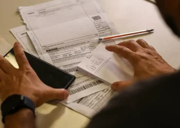 Pessoa organizando contas e segurando smartphone sobre mesa com papéis e caneta