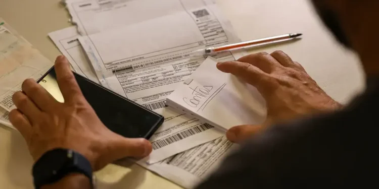 Pessoa organizando contas e segurando smartphone sobre mesa com papéis e caneta