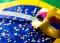 Máscara dourada decorada com detalhes em rosa e glitter sobre bandeira do Brasil decorada com confetes coloridos