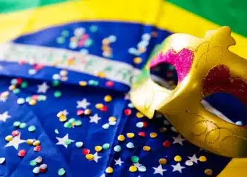 Máscara dourada decorada com detalhes em rosa e glitter sobre bandeira do Brasil decorada com confetes coloridos