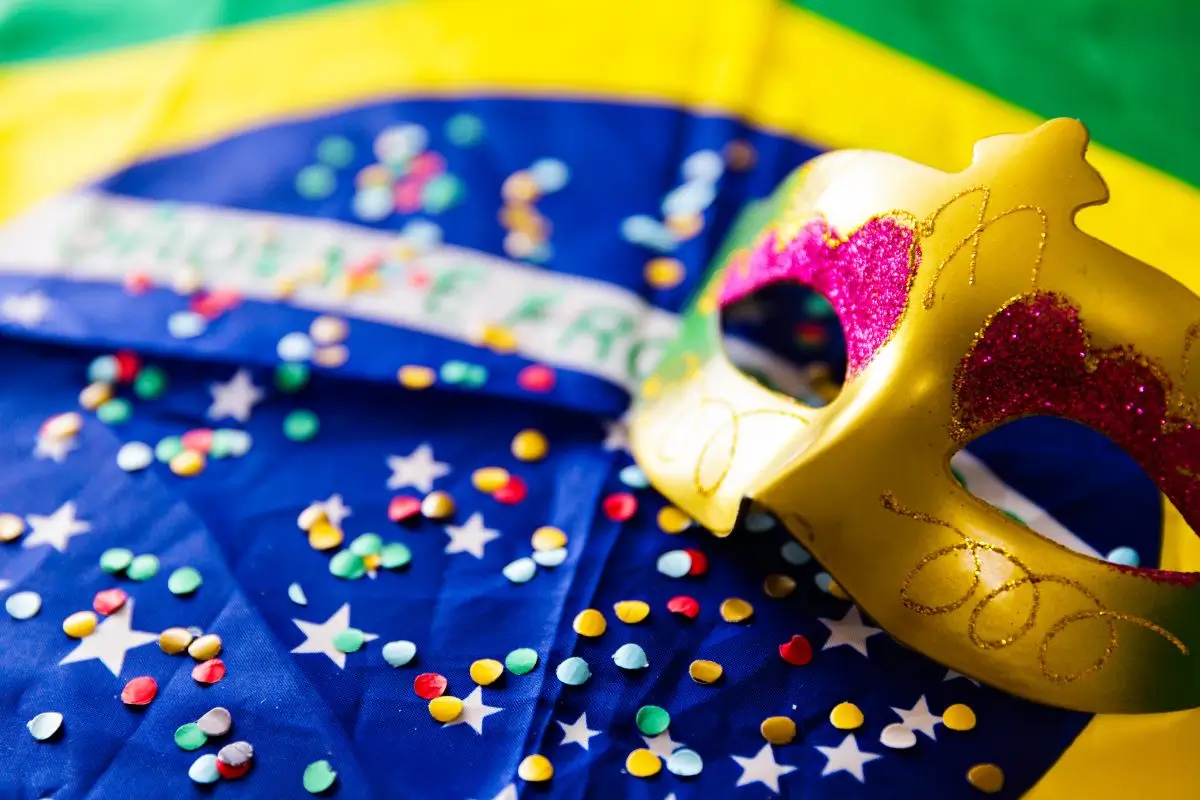Máscara dourada com glitter sobre a bandeira do Brasil decorada com confetes, simbolizando as celebrações do Carnaval 2026.