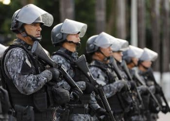 Policiais militares alinhados usando uniforme camuflado, capacete com visor e armamento tático.
