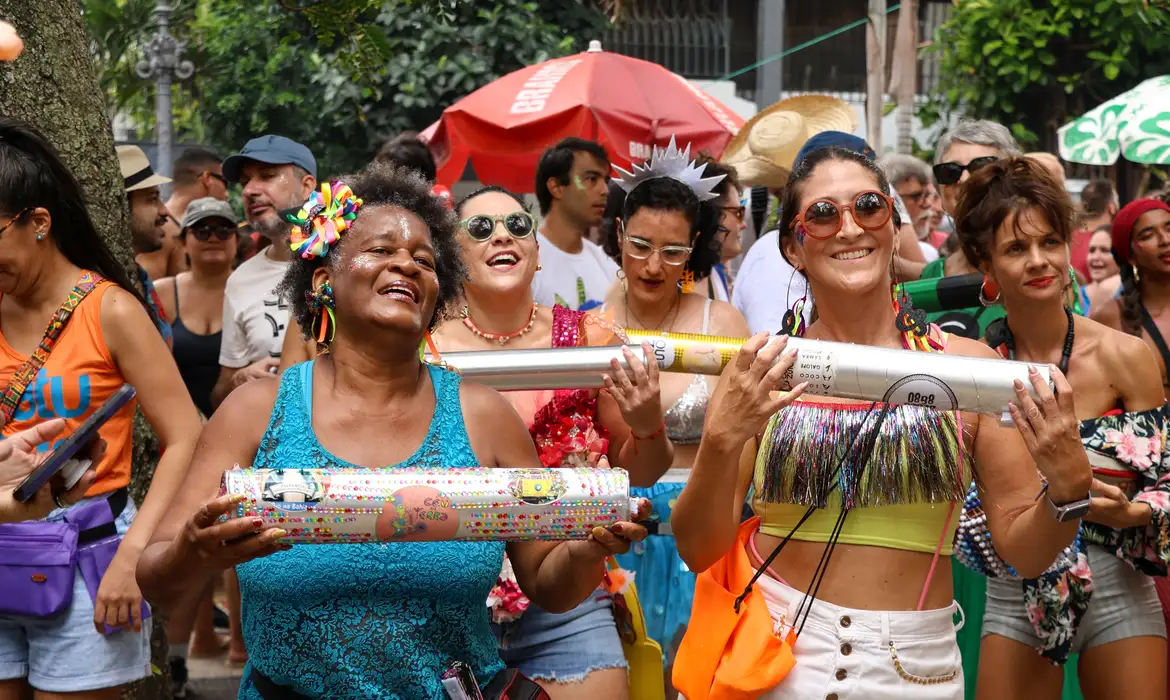 Mulheres alegres participam de bloco de Carnaval ao ar livre Mulheres sorrindo e tocando instrumentos coloridos durante bloco de Carnaval festivo na rua