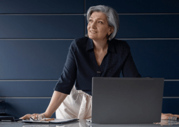 Mulher madura com cabelos grisalhos olhando para cima, ao lado de laptop em mesa clara com parede azul ao fundo