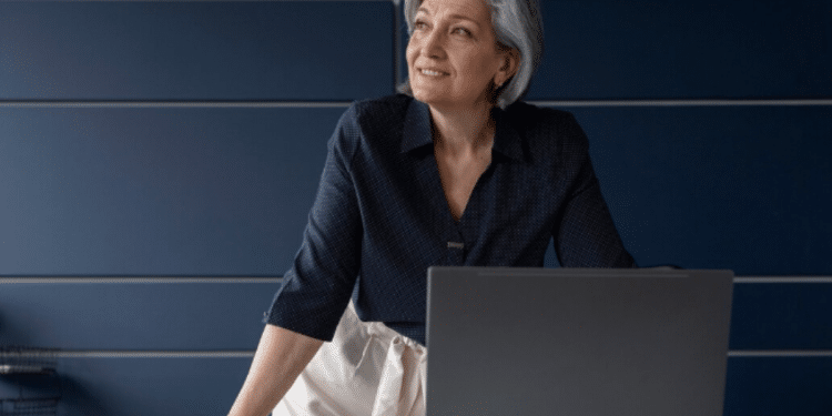 Mulher madura com cabelos grisalhos olhando para cima, ao lado de laptop em mesa clara com parede azul ao fundo