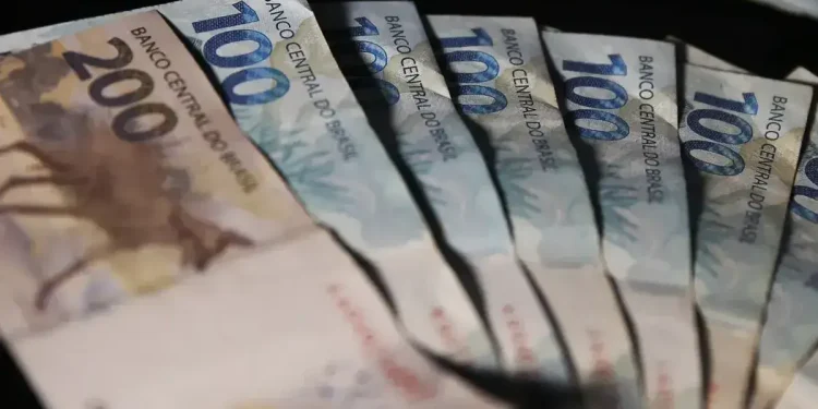 Cédulas de cem e duzentos reais dispostas em leque sobre fundo escuro.