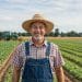 Agricultor sorridente usando chapéu de palha e jardineira em um campo cultivado, com um trator ao fundo.