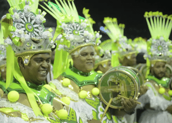 Passistas em desfile de carnaval com fantasias verde e prata, representando as festividades de 2026