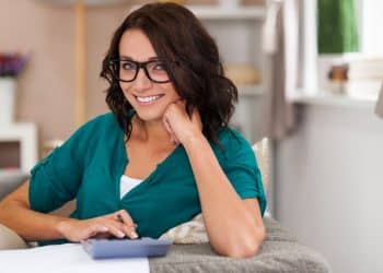 Mulher sorridente de óculos e camisa verde usando calculadora em ambiente doméstico aconchegante.