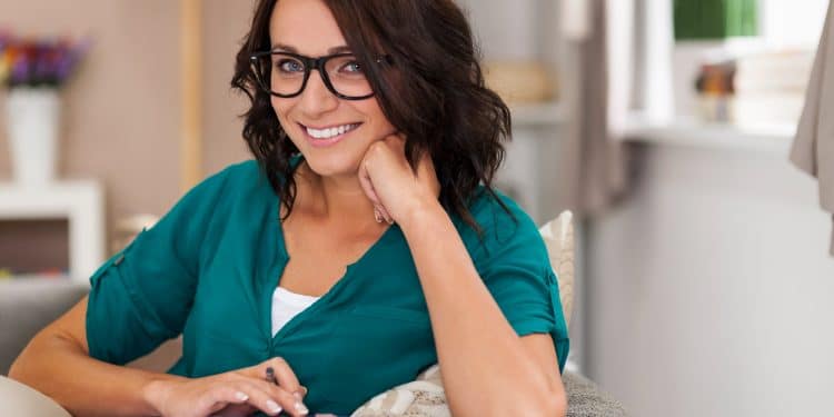 Mulher sorridente de óculos e camisa verde usando calculadora em ambiente doméstico aconchegante.