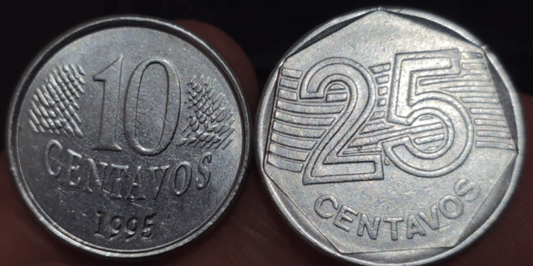 Grande quantidade de moedas de Real em destaque, com foco numa moeda de 25 centavos prateada ao centro e moedas de 1 Real douradas ao redor, evidenciando detalhes de relevo.
