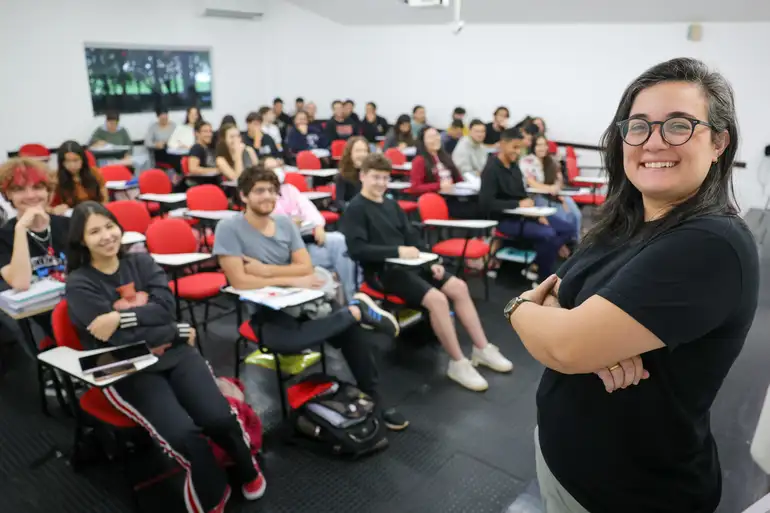 Professora sorridente à frente de uma sala cheia de alunos atentos em carteiras vermelhas