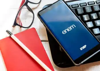 Celular com tela do aplicativo ENEM, óculos, caderno vermelho e lápis sobre mesa branca