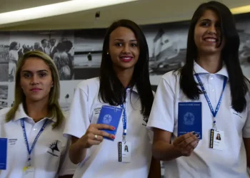 Três jovens sorridentes segurando carteira de trabalho azul e usando uniforme branco de trabalho