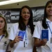 Três jovens sorridentes segurando carteira de trabalho azul e usando uniforme branco de trabalho