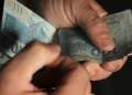 Mãos contando notas de cem reais simbolizando pagamento de benefício previdenciário reajustado.