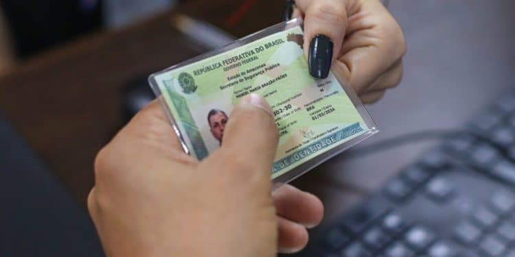 Mãos segurando a nova Carteira de Identidade Nacional plastificada com teclado de computador ao fundo