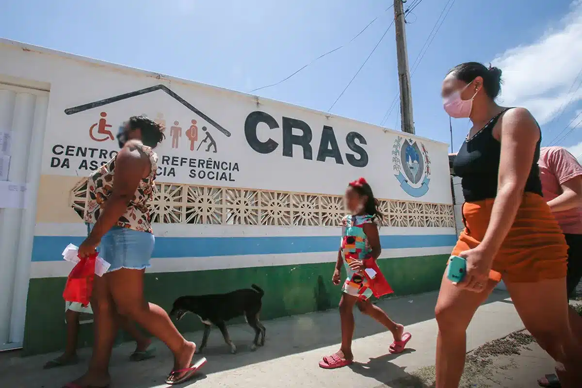 Pessoas caminhando em frente ao Centro de Referência de Assistência Social (CRAS), com mural de informações e o logo institucional ao fundo.