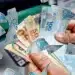 Cédulas de 100 e 50 reais sendo manuseadas, com notas de 100 reais flutuando, representando o impacto do reajuste do salário mínimo.
