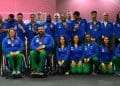 Grupo de atletas e paratletas da delegação brasileira vestindo uniformes oficiais azuis e verdes, alguns em cadeiras de rodas e outros em pé, exibindo suas medalhas de competição.
