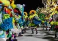 Integrantes de escola de samba desfilam no Carnaval 2026 com fantasias coloridas e bandeiras do Brasil.